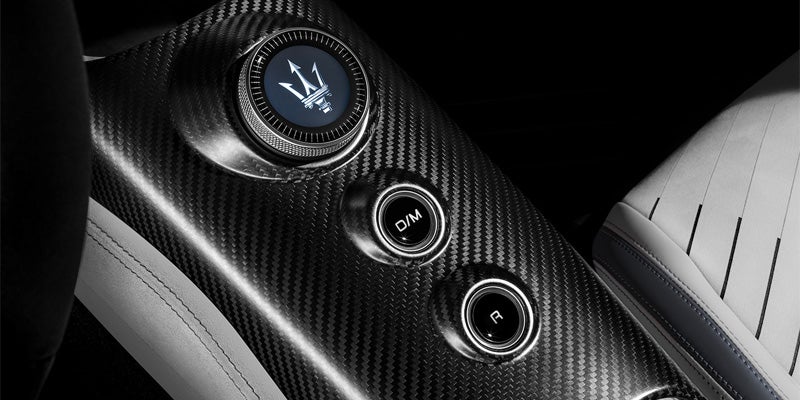 A close-up of a carbon fiber car console with a circular dial displaying the Maserati logo, and two round buttons labeled D/M and R.