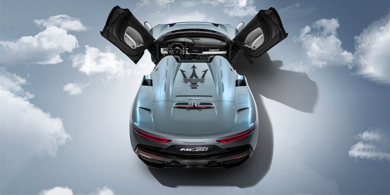 Top-down view of a light blue Maserati sports car with butterfly doors open upwards, against a cloudy sky background.