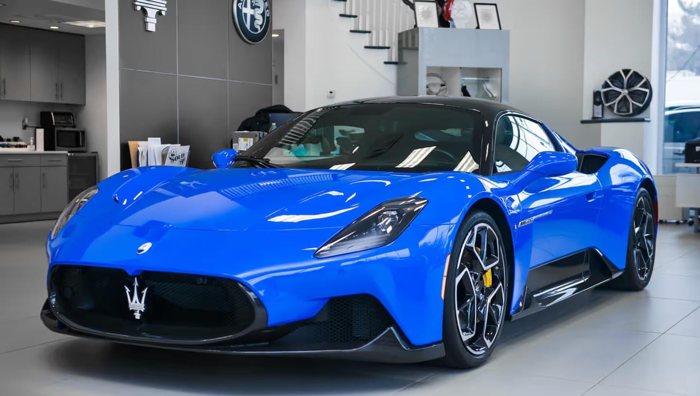 Front view of a shiny blue Maserati MC20 sports car inside a showroom with white and gray walls.