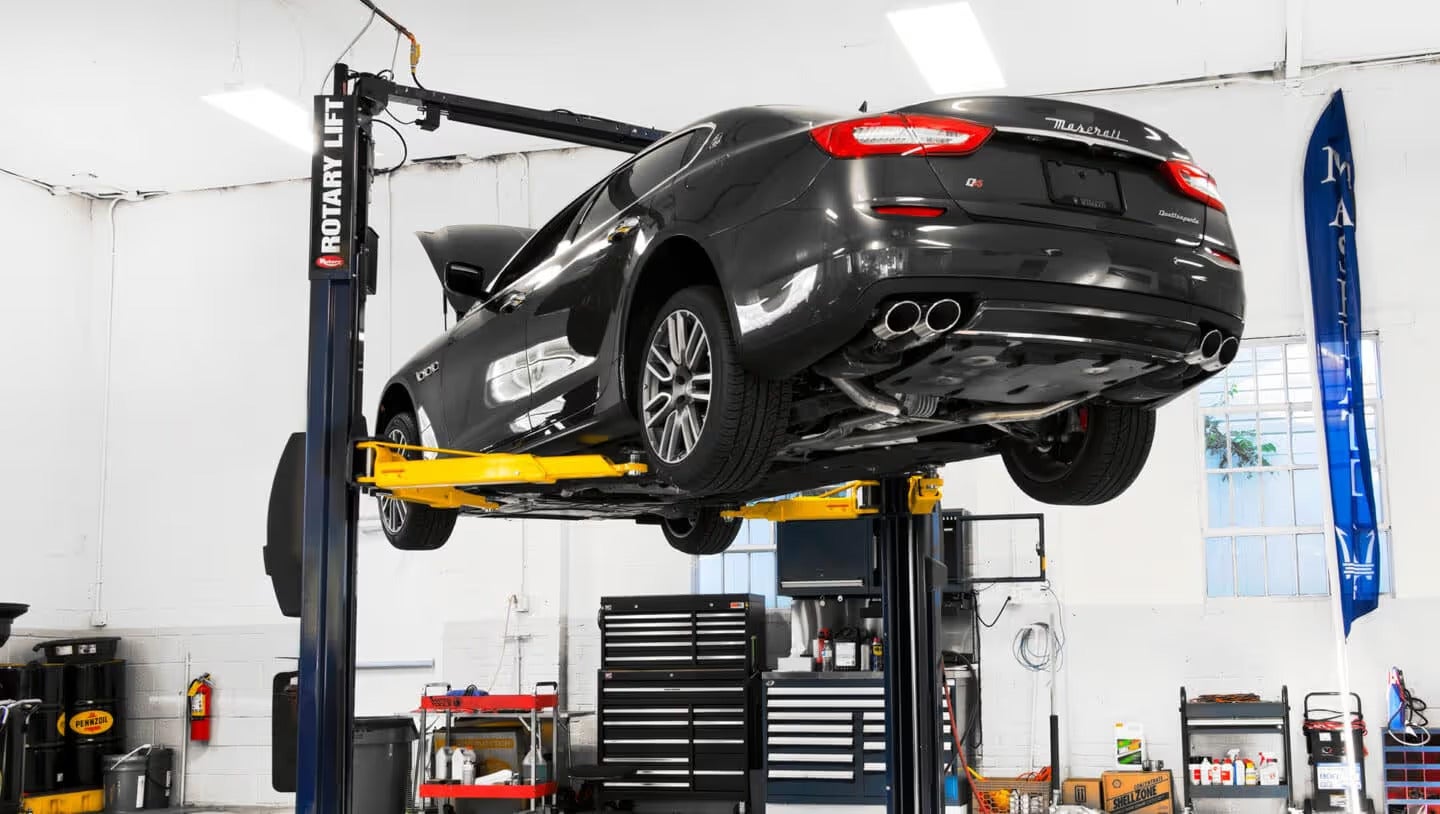 A black Maserati sedan is elevated on a yellow car lift inside a clean, white garage. Its hood is up.